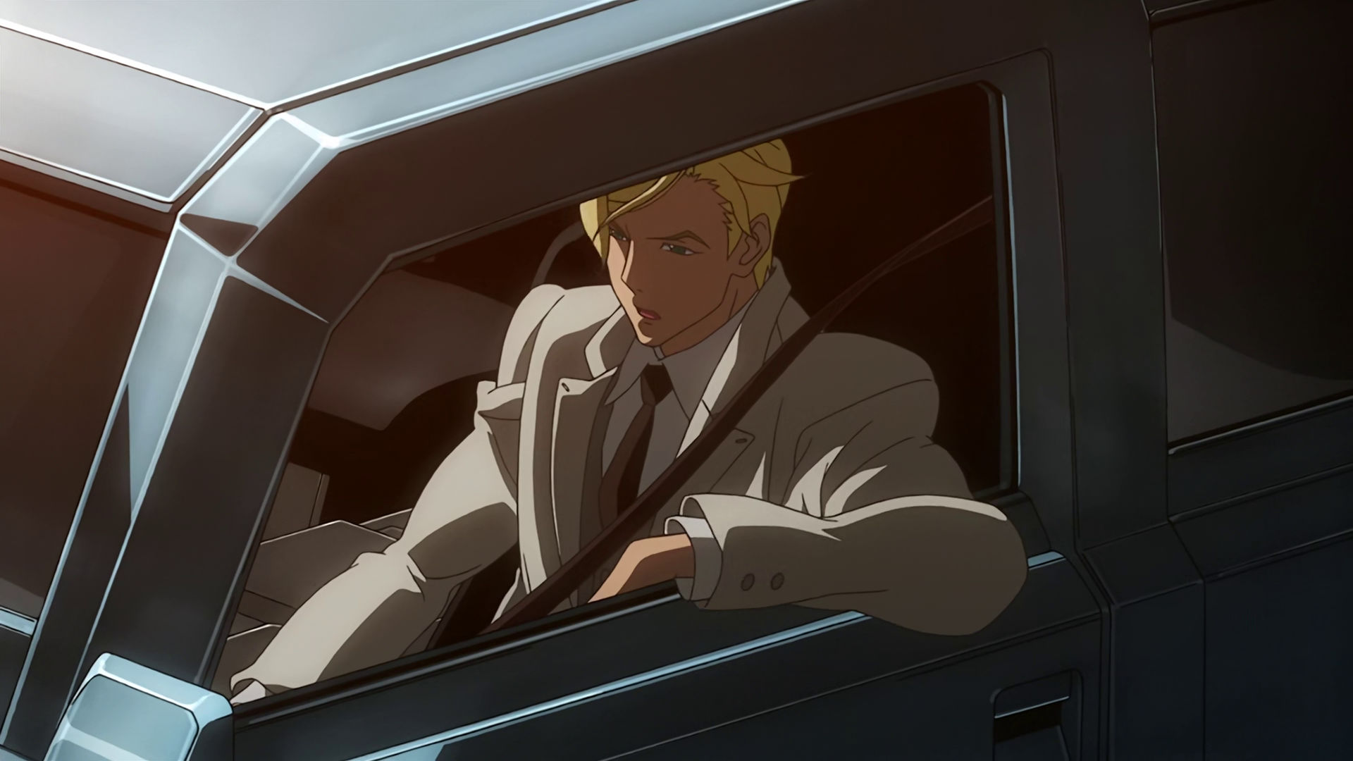 McGillis sitting inside a car, wearing an off-white suit and seatbelts, he looks serious.
